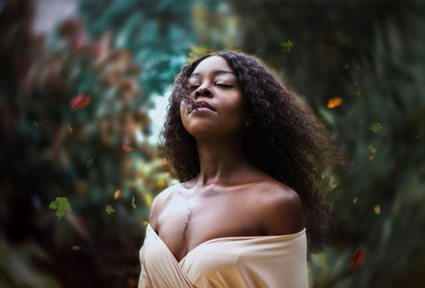 A middle-aged woman with dark skin and curly hair wearing an ivory top stands with trees blurred behind her.