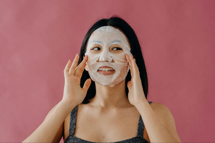 A woman smiles while pressing a sheet mask onto her face.