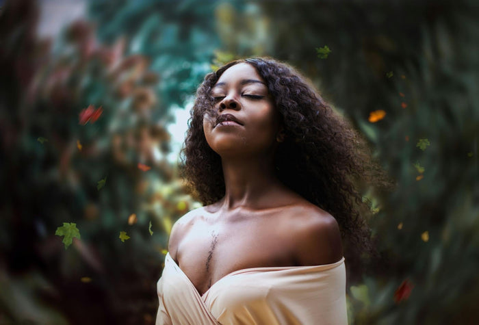 A middle-aged woman with dark skin and curly hair wearing an ivory top stands with trees blurred behind her.